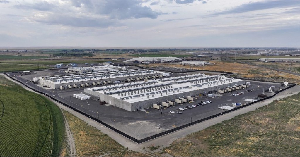 Aerial view of a large fenced data center campus: long gray buildings with dense rows of rooftop cooling equipment, parking and access roads inside the perimeter, green farmland on one side and open arid land on the other.